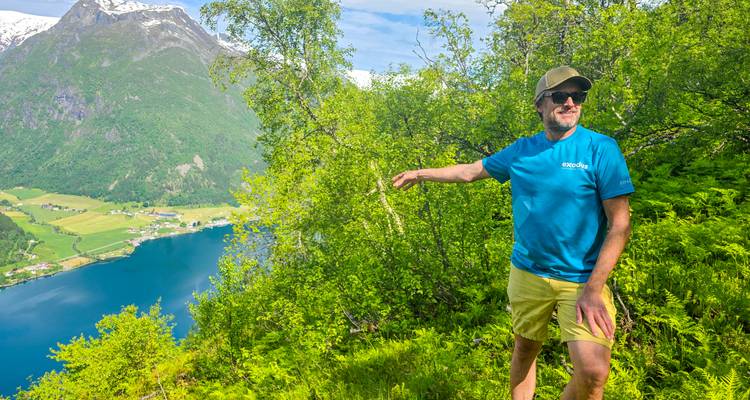Un guide fait un geste en direction d'une vue de fjord bordée d'arbres tout en se tenant au milieu d'une végétation luxuriante.