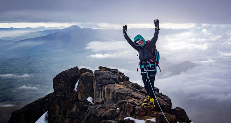 Escalador de pie triunfalmente en la cumbre rocosa sobre nubes ondulantes y picos brumosos