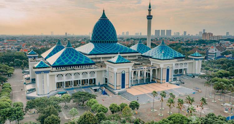 Vue aérienne de la vaste mosquée Masjid Al-Akbar au toit bleu entourée d'arbres et de l'horizon urbain.