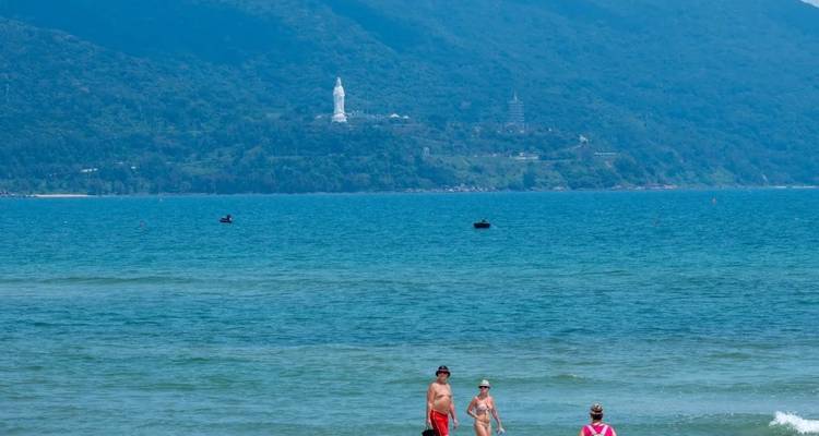 Strandszene mit Menschen, die im Meer waten, und einer Statue, die in der Ferne auf einem Hügel sichtbar ist.