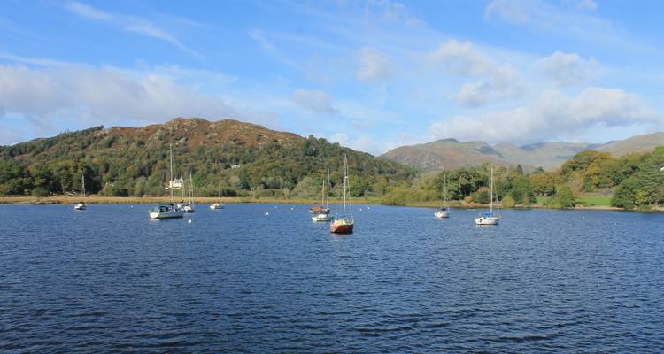 A tranquil lake dotted with sailboats is framed by forested hills and distant mountains under blue skies.