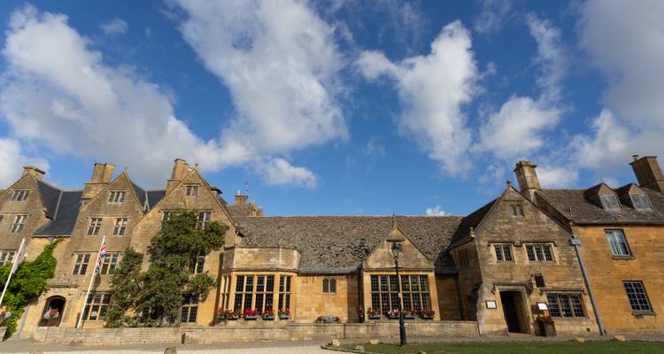 Wide-angle view of honey-colored stone buildings typical of an English Cotswolds village under a bright blue sky with scattered clouds.