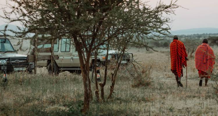 Two Maasai men in traditional shuka cloth walk across open savanna near safari vehicles at dawn.