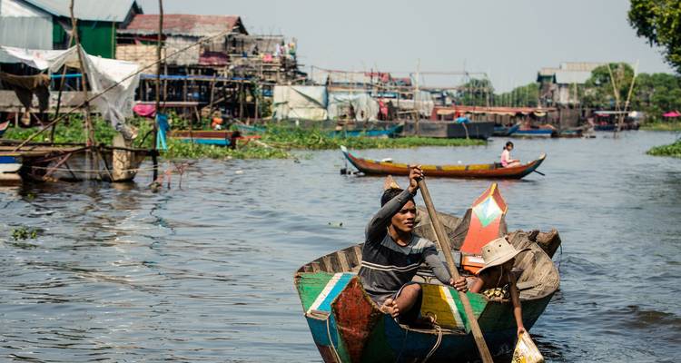 Lokale dorpelingen die in houten bootjes voorbij stelpenhuizen in een drijvend dorp op een rivier peddelen.