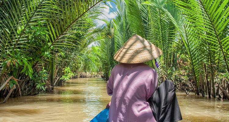 Vrouw in traditionele punthoed die met een klein bootje door een weelderig, met palmen omzoomde kanaal in de Mekongdelta peddelt.