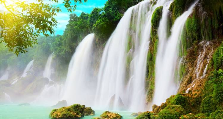 Powerful curtains of water cascade over moss-covered cliffs into a turquoise pool below a tropical forest canopy.