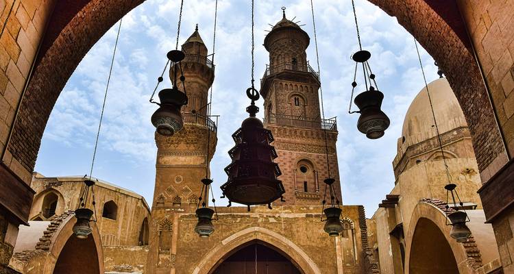 Looking up through an archway toward twin minarets of an old Cairo mosque framed by hanging lanterns against a partly cloudy sky.