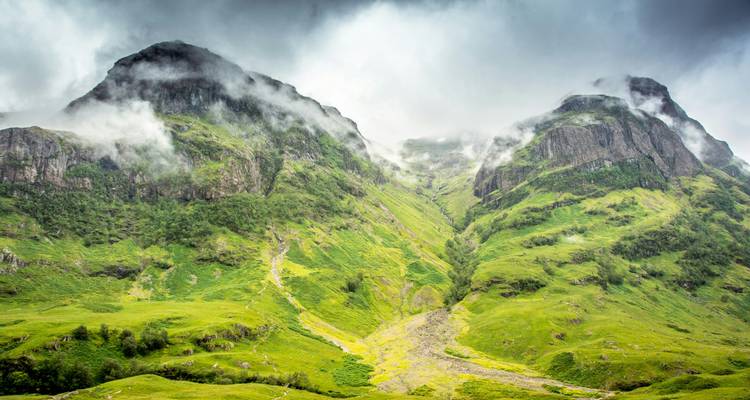 Nebelumhüllte grüne Berge von Glencoe mit tiefhängenden Wolken, die durch ein steiles Tal driften.