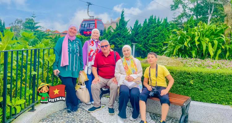 Travelers resting on a bench surrounded by lush landscaped gardens, with a red cable car passing overhead.