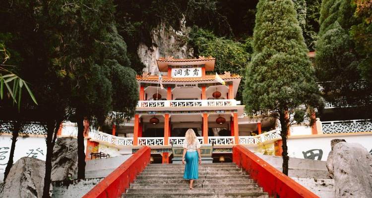 A woman in a turquoise dress climbs red stairs toward an ornate hill-side temple surrounded by jungle.