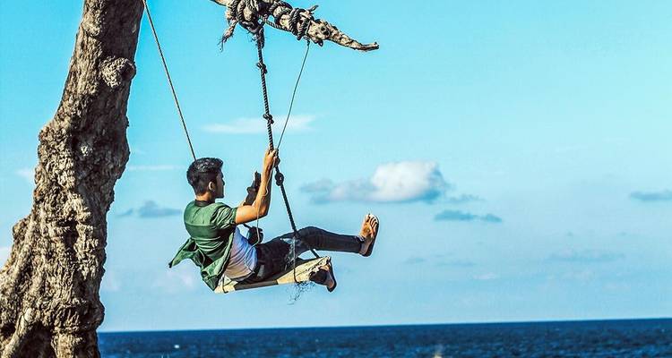 Young man swings from a rope tied to a seaside tree, high above a deep-blue ocean.