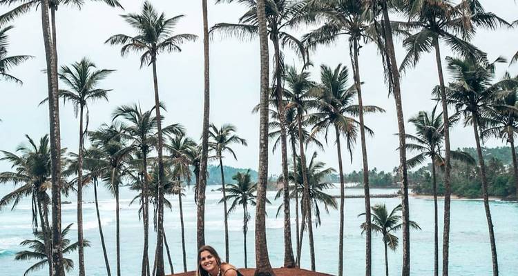 Woman among tall palm trees with ocean in the background.
