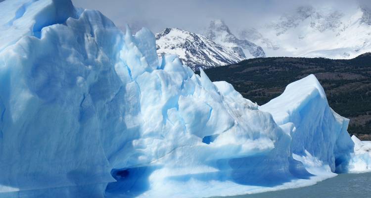 Ein massiver blauer Eisberg vor einer Kulisse von schneebedeckten Bergen.