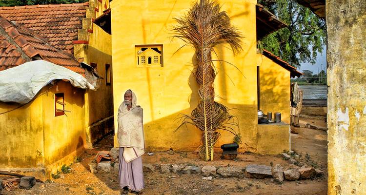 Ältere Person in einen Schal gehüllt, die vor gelb gestrichenen traditionellen Häusern steht.