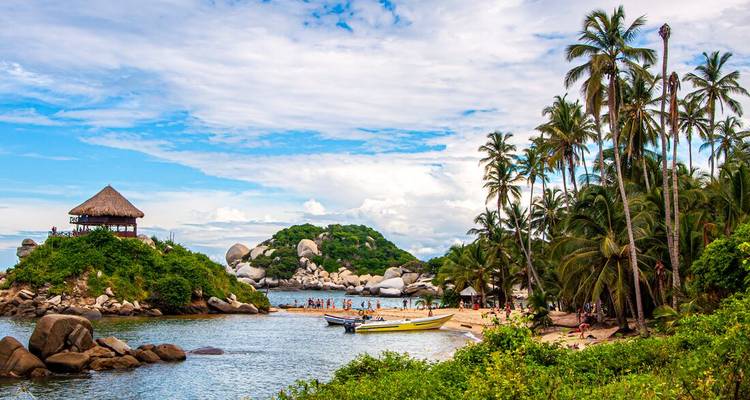 Baie tropicale avec plage bordée de palmiers, cabane d'observation rustique sur un îlot rocheux et visiteurs profitant de l'eau