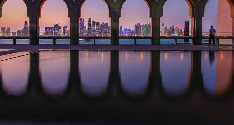 Horizonte de la ciudad de Doha al atardecer visto a través de arcos con reflejos en el agua.