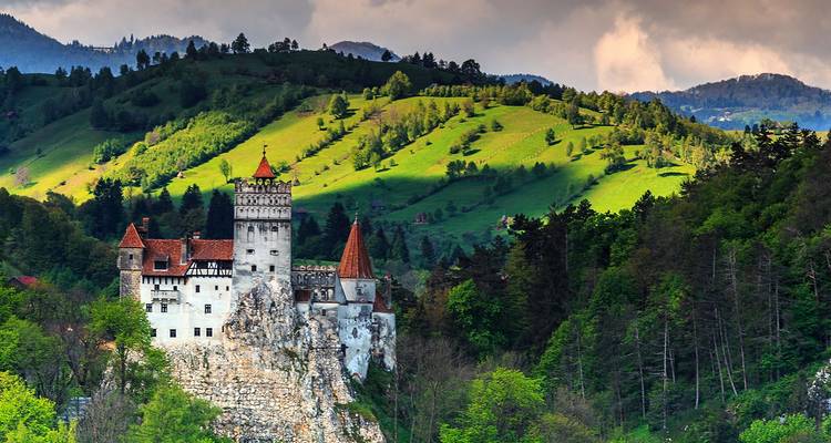 Bran Castle framed by lush green hills and a dramatic cloudy sky in bright daylight.