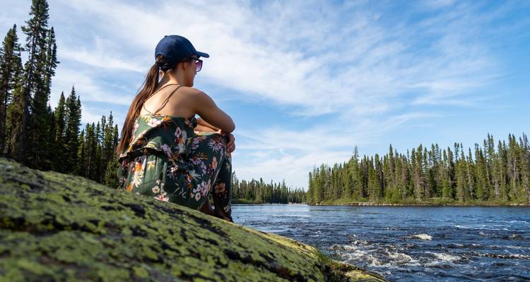 Una mujer con un vestido floral se sienta en una roca cubierta de musgo mirando un río rápido en el bosque bajo un cielo azul.