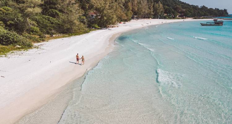 A couple walks hand-in-hand along a pristine white-sand beach beside clear turquoise water on a tropical island.