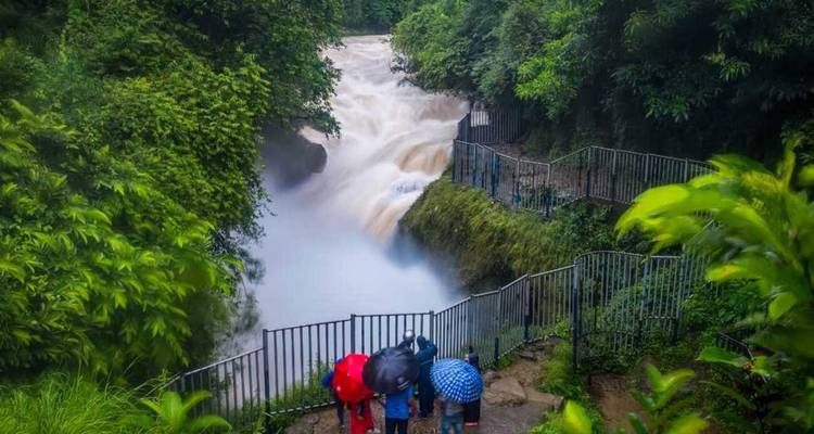 Waterfall surrounded by lush greenery, with people observing it from a fence.