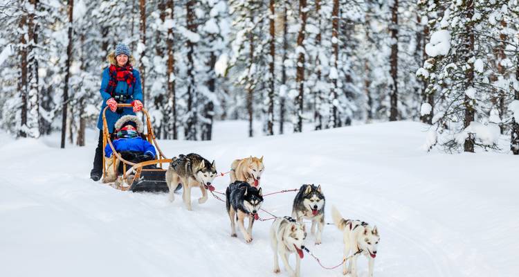 Une équipe de huskies tire un traîneau en bois avec des passagers chaudement vêtus à travers une forêt de pins chargée de neige.