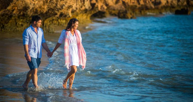 Romantic couple walk barefoot through gentle surf along a sunny turquoise beach beside cliffs.