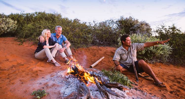 Small group relaxes around a crackling campfire on red sand while a guide gestures to the horizon.