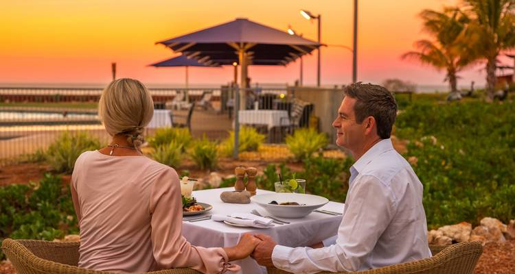 Couple enjoys a candle-lit seaside dinner at sunset with palm trees and umbrellas behind them.