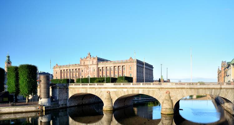 Het Zweeds Parlamentsgebouw torens uit boven een bogen stenen brug onder een heldere blauwe lucht.