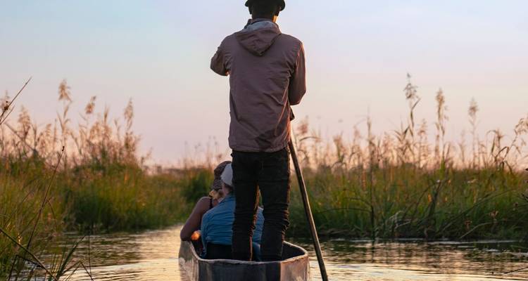Guide poles a dugout canoe through tall reeds with two travelers seated at dusk