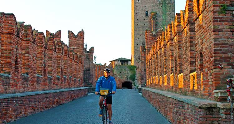 Un ciclista cruza el puente medieval de ladrillo de Verona al amanecer con luz cálida sobre las murallas almenadas.
