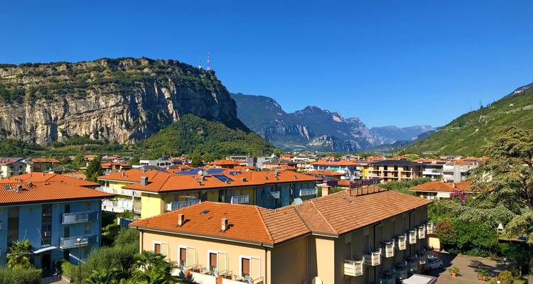 Vista sobre un pueblo de techos de terracota situado contra acantilados verdes empinados y picos lejanos de los Dolomitas bajo un cielo despejado.