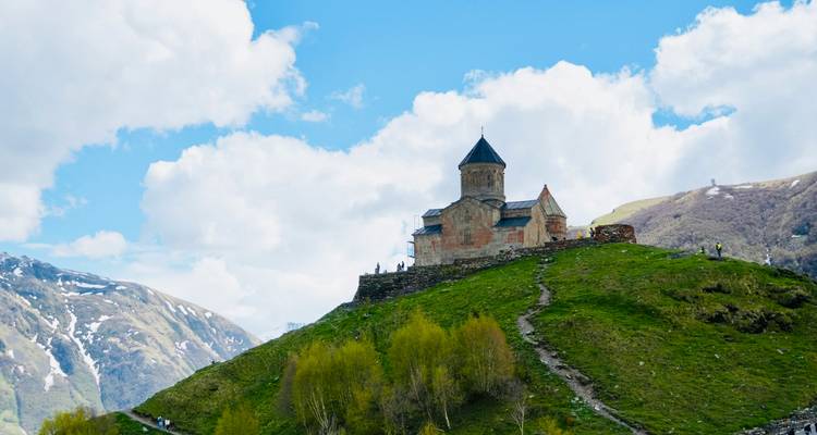 Historische kerk op een heuvel met mensen eromheen en bergachtige achtergrond.