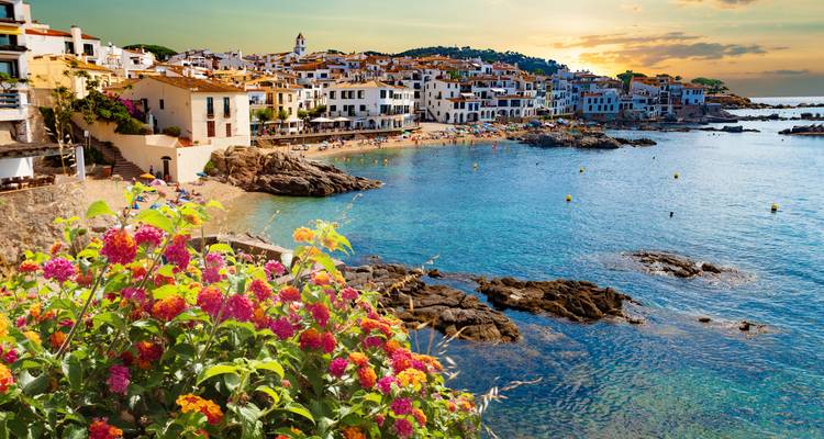 Schilderachtig mediterraan dorpje aan een turkooizen baai tijdens het gouden uur met kleurige bloemen op de voorgrond.