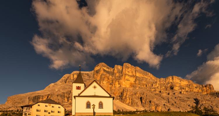 A picturesque chapel in front of a dramatic mountain range under a partly cloudy sky.
