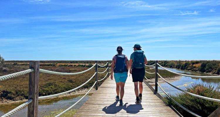 Zwei Rucksacktouristen überqueren einen hölzernen Steg über Küstenfeuchtgebiete in Richtung eines weiten Horizonts.