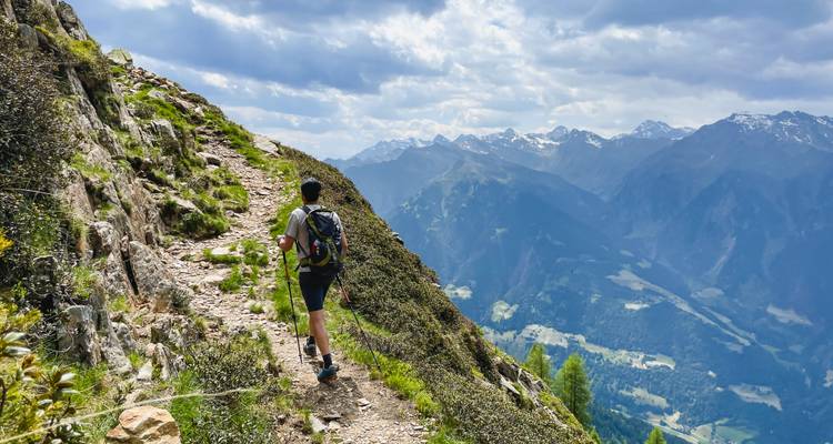 Hiker strides along a narrow rocky trail high above a dramatic valley.