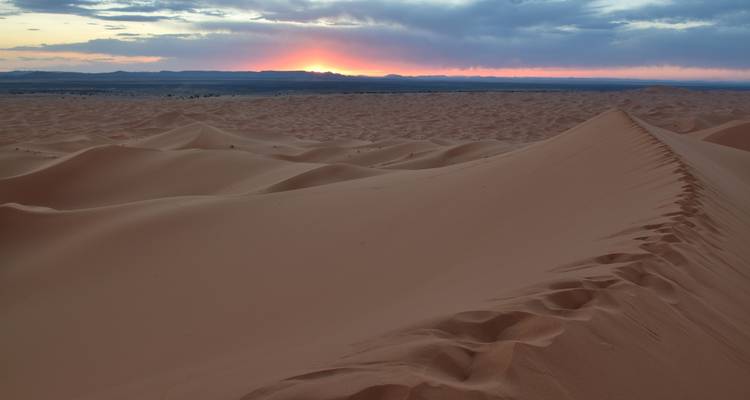 Desert landscape at sunset with rolling sand dunes under a colorful sky.
