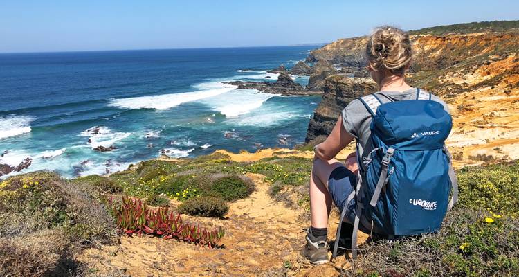 Junge Wanderin mit großem Rucksack sitzt auf einem Küstenweg mit Blick auf dramatische Klippen und türkisfarbene Wellen.
