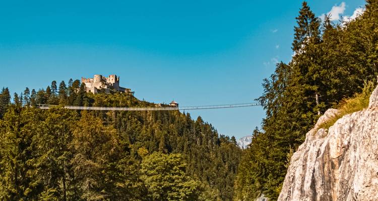 Eine Hängebrücke, die zu einer historischen Burg auf einem bewaldeten Hügel führt.