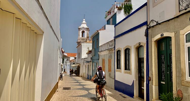 Un cycliste traversant une rue pavée étroite bordée de maisons colorées de l'Algarve et d'un clocher distant.