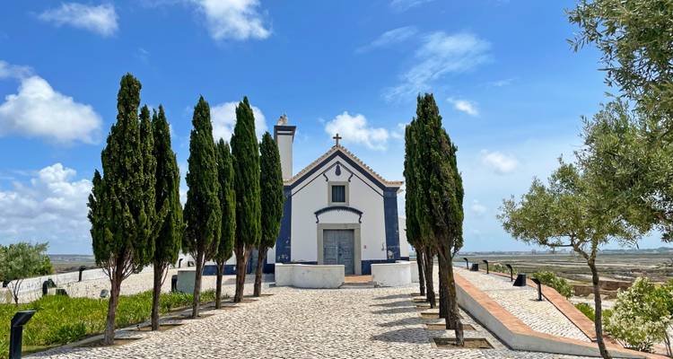 Capilla enmarcada por altos cipreses al final de un sendero empedrado con vista a salinas bajo un cielo azul.