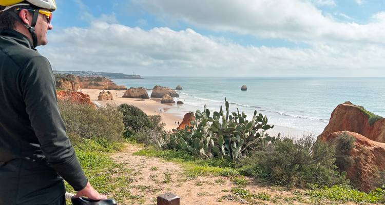 Ciclista con casco hace una pausa en un sendero en la cima de un acantilado con vista a playas doradas, pilas marinas y océano en calma.