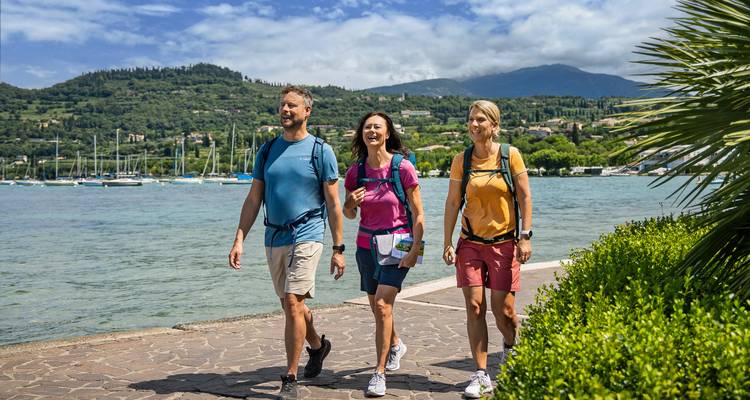 Trois amis se promènent le long du lac de Garde avec des voiliers et des montagnes boisées en arrière-plan.