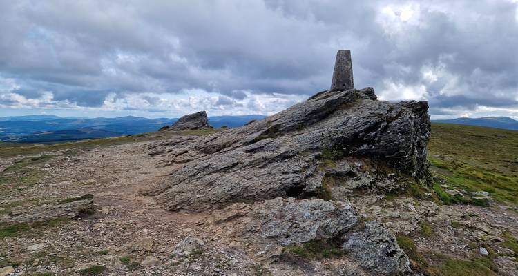 Sommet rocheux avec pilier de triangulation en béton dominant des collines ondulantes sous des nuages spectaculaires.