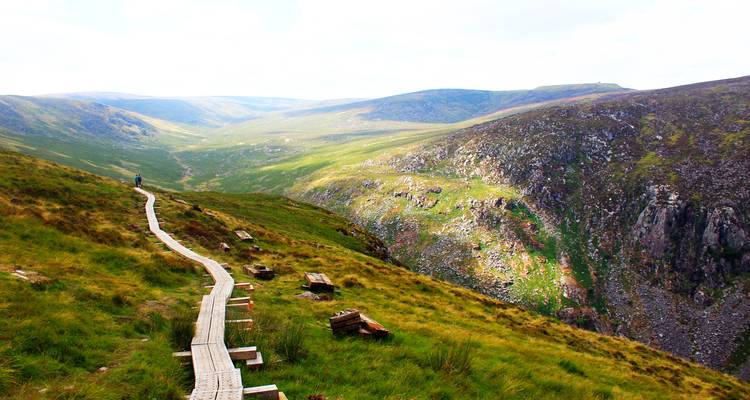 Sentier de promenade en bois s'étendant à travers la vallée verte des montagnes de Wicklow avec un randonneur solitaire au loin.