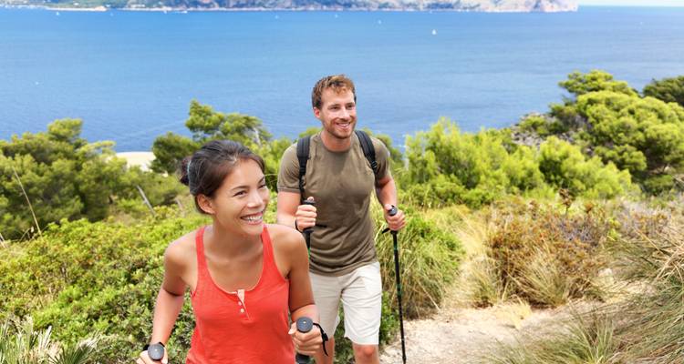 Two hikers smiling and walking with trekking poles along a sea view trail.