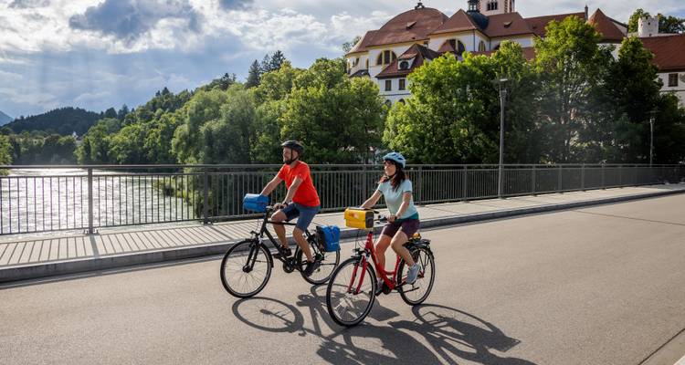Ciclistas cruzando un puente sobre el río con la luz del sol brillando en el agua y un edificio histórico detrás.