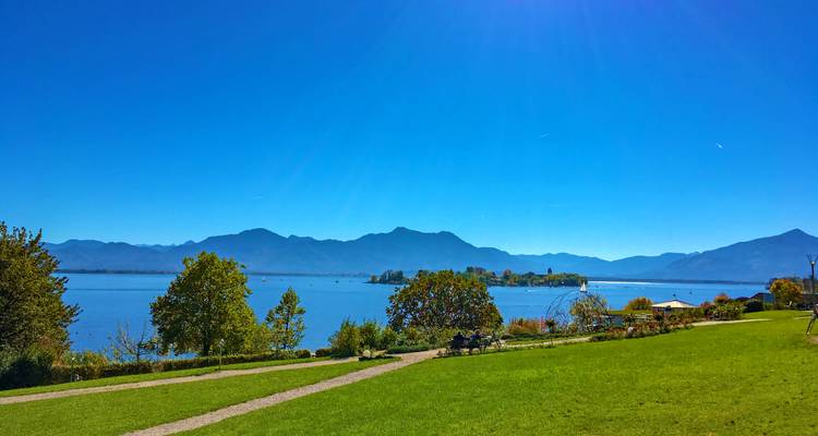 Amplio césped junto al lago con vista a un pueblo isleño y cadena montañosa bajo cielos azules despejados.