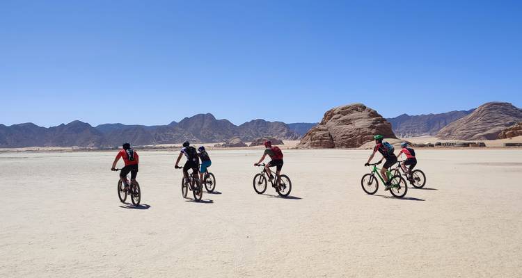 Group of cyclists riding in a desert with rocky formations in the background.